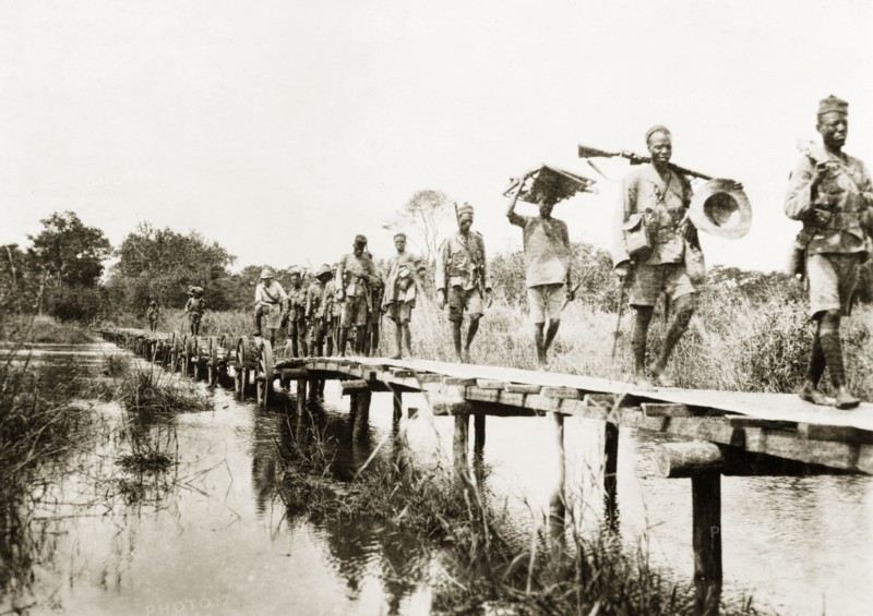 African schutztruppes on the march. A regiment of African schutztruppes march in single file across a wooden bridge over a river in German East Africa. German East Africa (Tanzania), circa 1910. Tanzania, Eastern Africa, Africa.