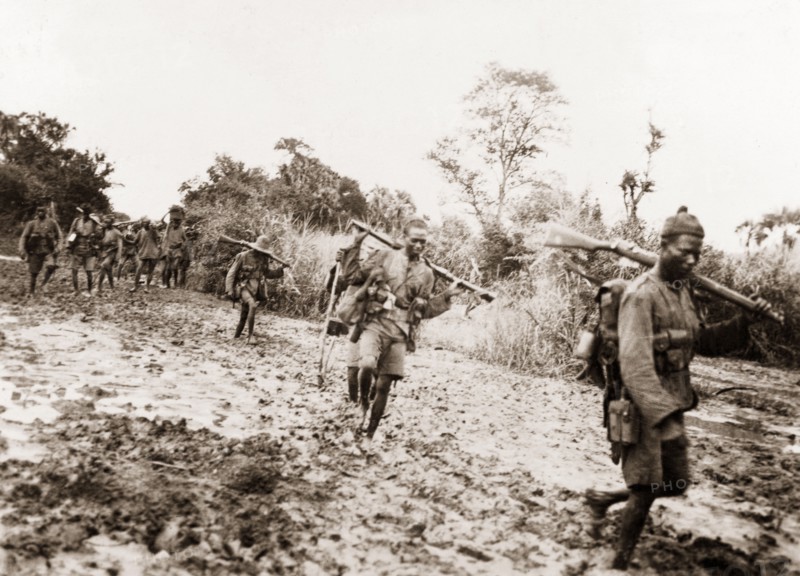 African schutztruppes on the march. A regiment of African schutztruppes trek across boggy land, carrying their rifles slung over their shoulders. German East Africa (Eastern Africa), circa 1910., Eastern Africa, Africa.