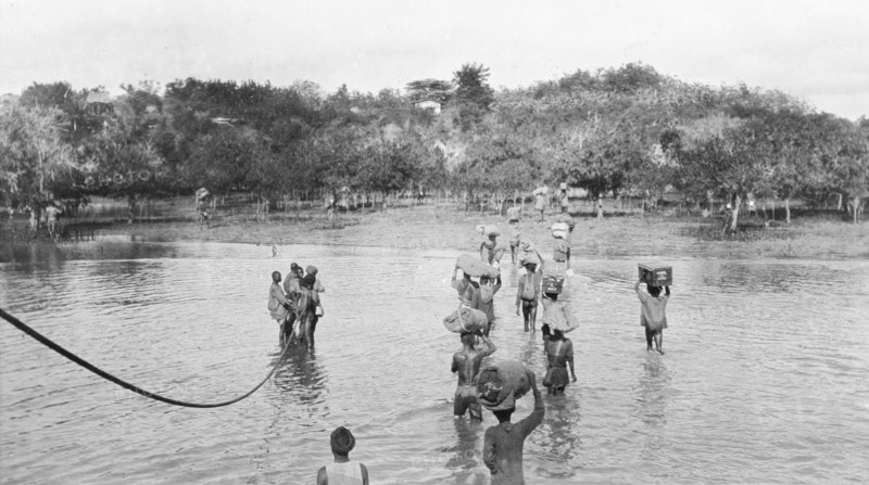 Disembarkation at Tanga'. African men disembark from a British boat during the Battle of Tanga, balancing bundles and trunks on their heads as they wade ashore through shallow water. An original caption comments that the men were carrying "the stores and equipment of a field ambulance" and refers to Captain Kennedy of the Indian Medical Service. Tanga, German East Africa (Tanzania), 3-5 November 1914. Tanga, Tanga, Tanzania, Eastern Africa, Africa.