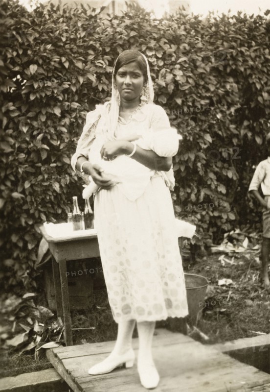 Indo-Trinidadian woman with baby. Portrait of an Indo-Trinidadian woman holding a baby in the crook of her arm. She wears a Western-style dress and shoes, with a traditional Indian 'dupatta' (head scarf). Siparia, Trinidad, circa 1931. Siparia, Trinidad and Tobago, Trinidad and Tobago, Caribbean, North America .