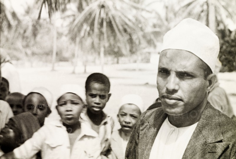 A Surahi type'. A Muslim man (possibly a teacher) stands in front of a group of children, staring into the camera. He wears a white kufi (cap) and is identified in a related caption as a 'Surahi type'. Possibly Mombasa, Kenya, 1947. Mombasa, Coast, Kenya, Eastern Africa, Africa.