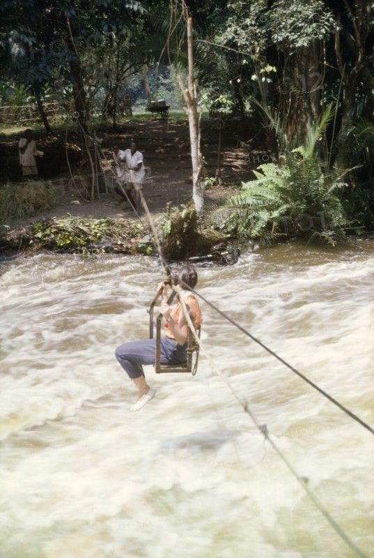 Elisabeth Lang Brown crosses to Kyantsore Island. Elisabeth, the wife of District Forestry Officer James Lang Brown, crosses the rapids of Kagera River by way of a cable chair lift. A related caption comments that she was crossing to Kyantsore Island to visit the Countess Antoinette Nuti (Toni), a colonial settler who had taken a lease on the island from the Omukama (King) on Ankole. Ankole, West Uganda, November 1963., West (Uganda), Uganda, Eastern Africa, Africa.