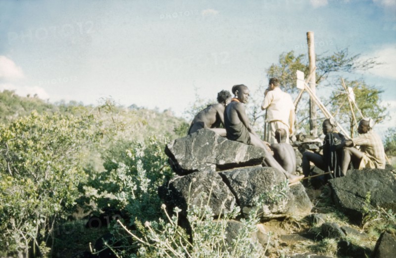 Henry Osmaston takes a bearing'. Accompanied by Suk porters, Henry Osmaston (Working Plans Officer of the Forest Department) stands at the base of a 'kituti' (cairn) marked with bamboo sticks, surveying Pcholio Peak in the Karasuk hills. Part of a forestry survey team, he uses a theodolite to locate and measure the distance to the next 'kituti'. Kenya, May 1959., East (Uganda), Uganda, Eastern Africa, Africa.