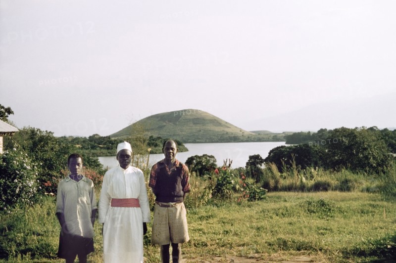 Portrait of three Ugandan house staff. Three Ugandan house staff on the shores of a lake. They are identified (left to right) as: "The toto, who did all the dirty work and cut the grass, ...Asanasio who waited at table and was (a) safari boy, and ...the cook Nikimia". Probably Saka, West Uganda, 1955., West (Uganda), Uganda, Eastern Africa, Africa.
