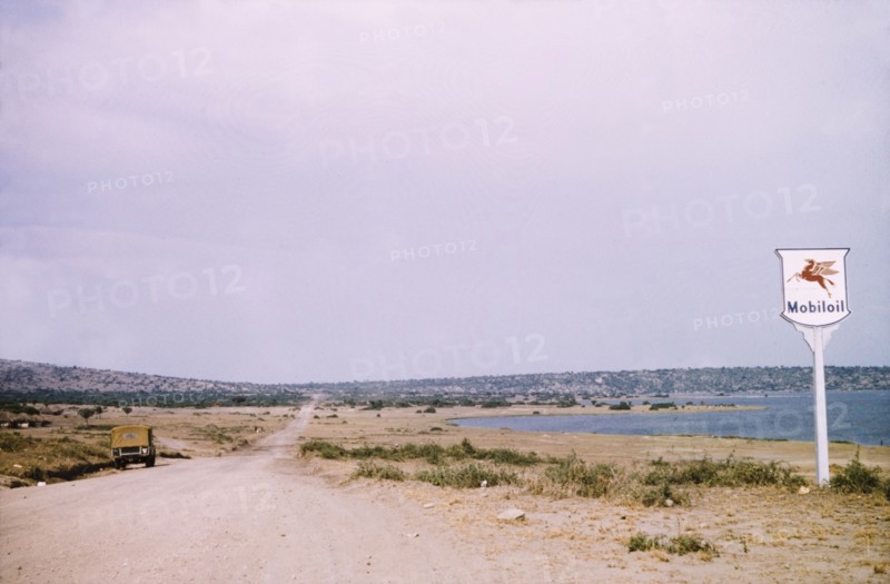 A rural road in Katwe. A Mobiloil sign stands beside a rural road in Katwe to Fort Portal, a fishing village located in the Queen Elizabeth National Park. Katwe's volcanic salt lake is visible in the distance. Katwe, West Uganda, August 1956., West (Uganda), Uganda, Eastern Africa, Africa.