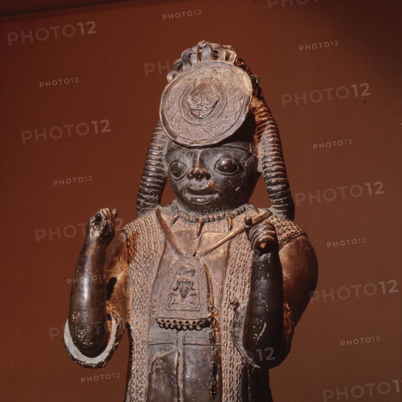 A large figure, one of the Tsoede bronzes, wearing an outer garment covered with cowrie shells, a necklace of leopards teeth and a pectoral with representations of a ram and birds