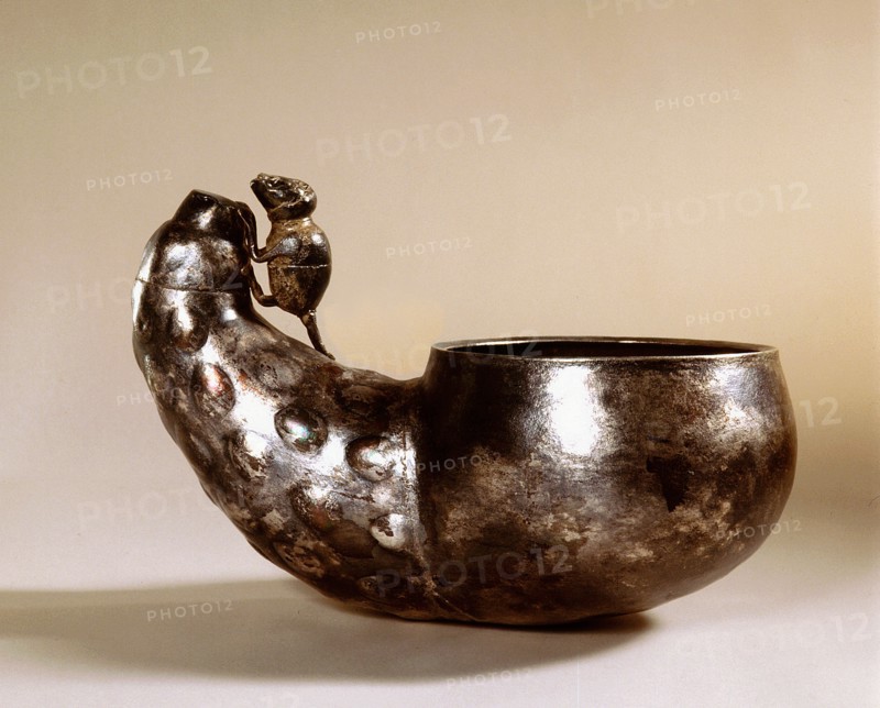 A cup made of beaten metal with a handle in the form of a mouse climbing over a vegetable
