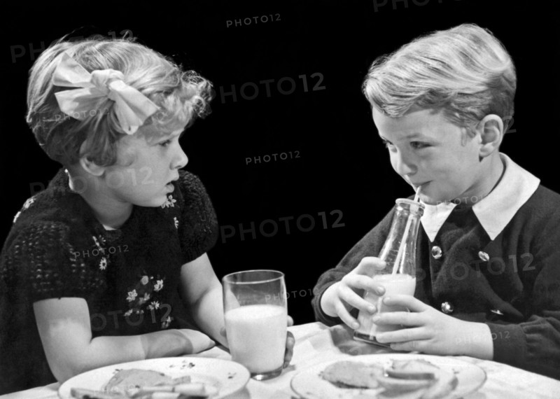 United States:  c. 1940.
A young boy and girl looking at each other as they drink milk at the lunch table.