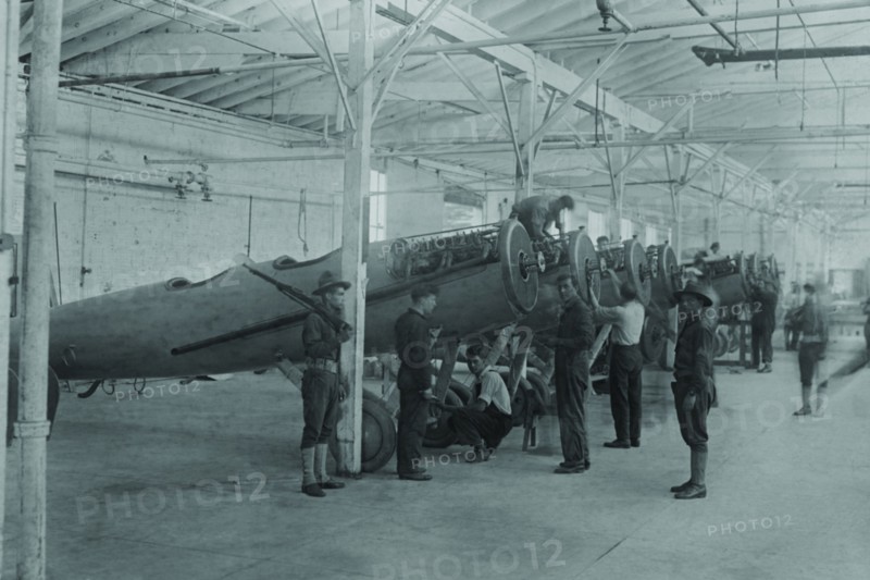 A line of airplanes is being assembled under the watchful eye of a gun-toting soldier as they are being completed to be sent to the European war; the factory is at College Point, Queens 1917