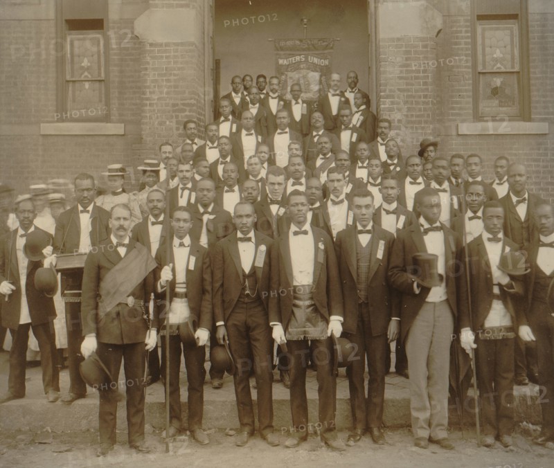 African American men posed at entrance to building, some with derbies and top hats, and banner labeled "Waiters Union" in Georgia 1899