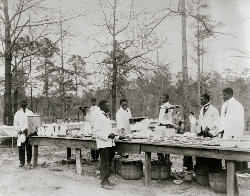 African American men preparing to serve a meal in an outdoor setting among trees 1900