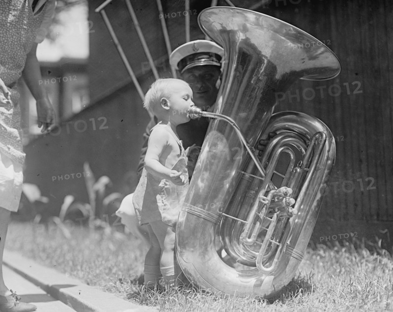 An nearly naked infant toots a tuba many times his size 1917