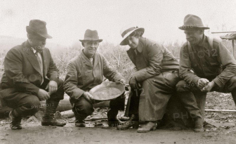 Group portrait of a woman and three men, crouched outside, smiling; one of the men is displaying a dish of panned gold 1910