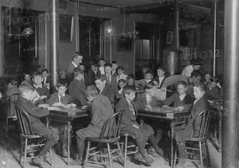 In the Newsboys Reading Room. Boys seated at tables playing gamers. 1909