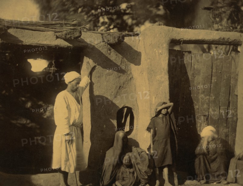 Man, woman, and children outside earth and wood structure 1880