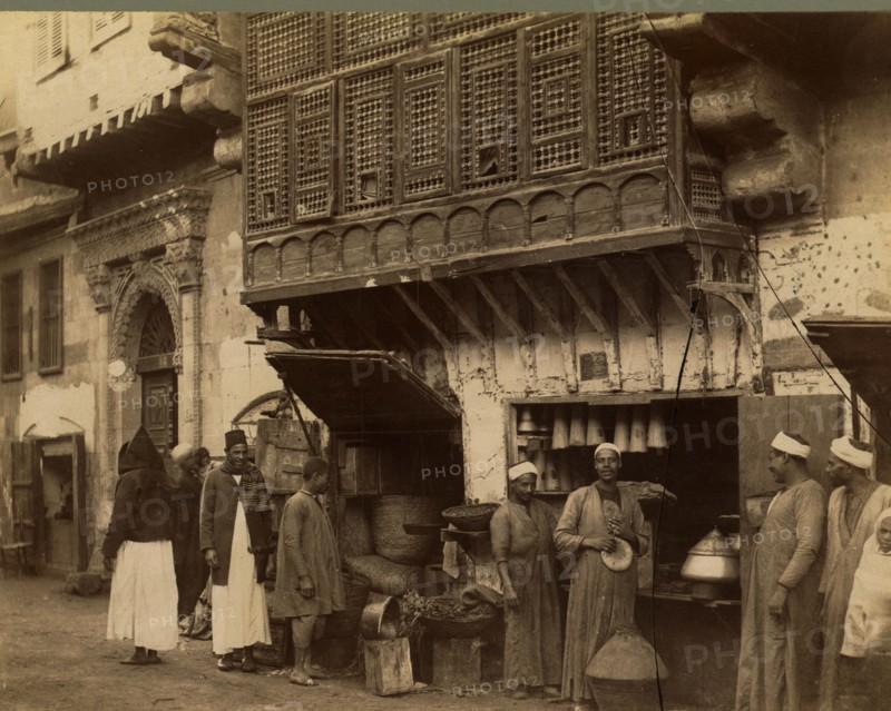 Men standing next to shop stall open to street, 1870