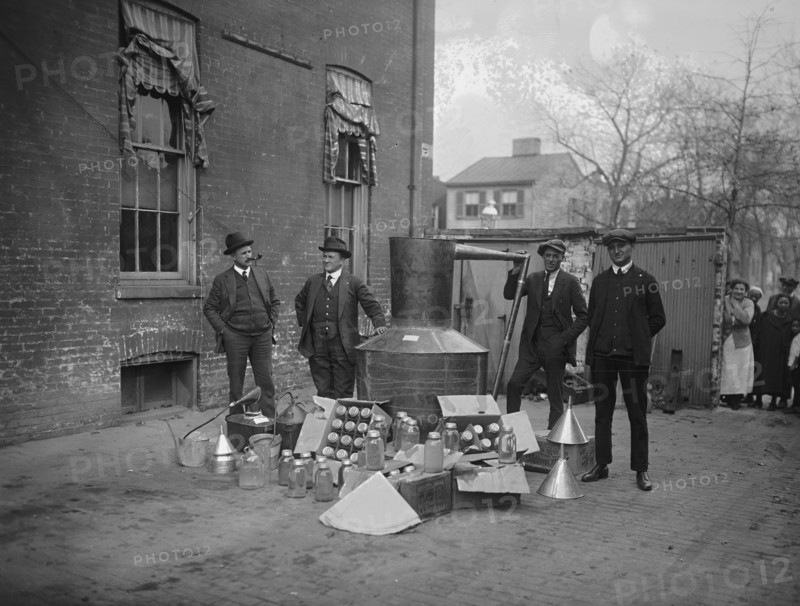 Onlookers watch as suited men stand in front of a large copper kettle still for making illegal liquor with boxes of bottles and funnels spread before them all for the manufacture of booze.
