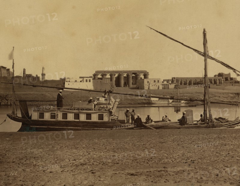 Several men on boat docked at water's edge, temple ruins of ancient Thebes in background, Luxor, Egypt. 1880