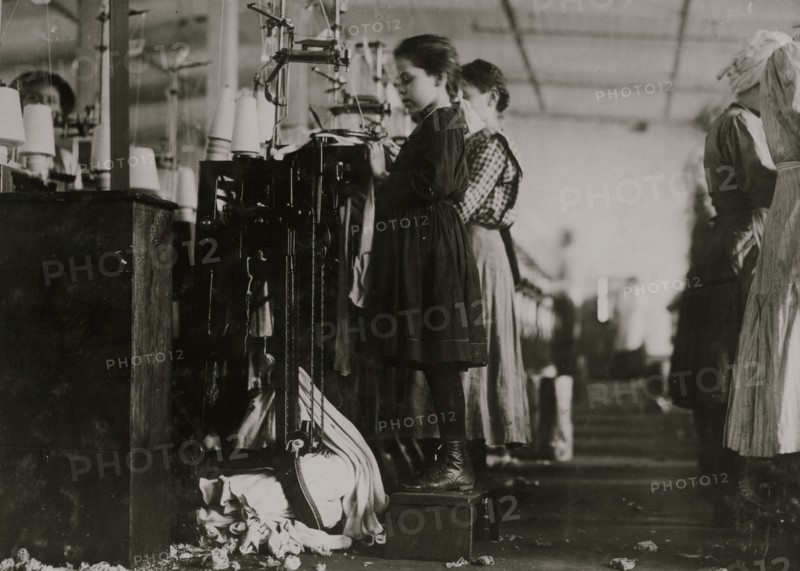 Knitter is a little girl so small she has to stand on a box to reach her machine. 1910