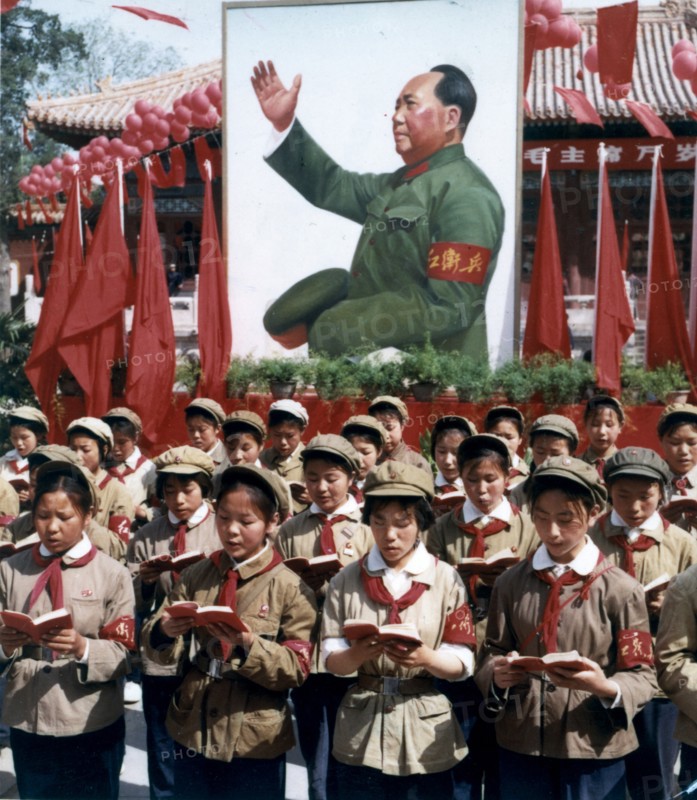 Children reading from mao's little red book at a rally with a portrait of mao in the background.