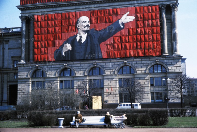A banner of lenin on the side of a building overlooking two elderly women sitting on a park bench in leningrad, ussr, 1960s.