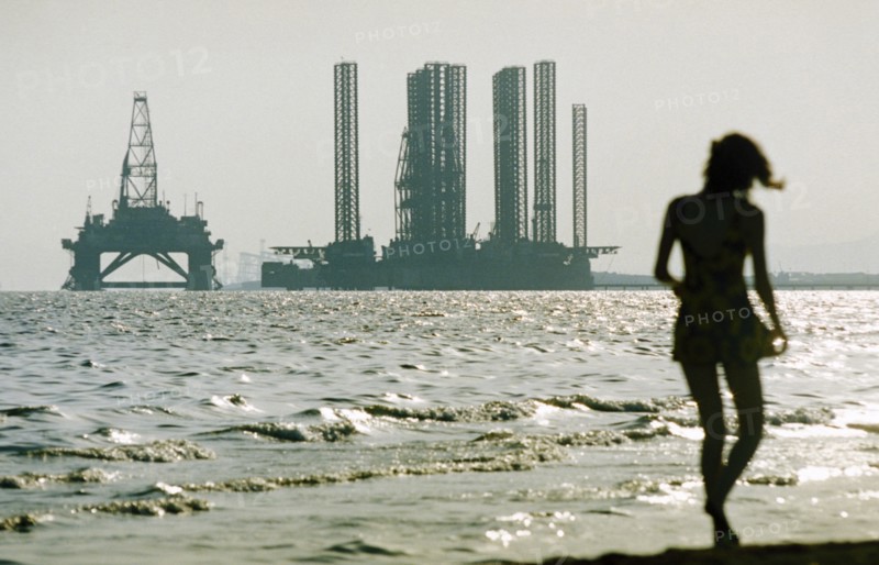 A woman in silhouette walking along shikhov beach on the shore of the caspian sea with an off-shore oil rig under construction in the background, baku, azerbaijan, october 2002.
