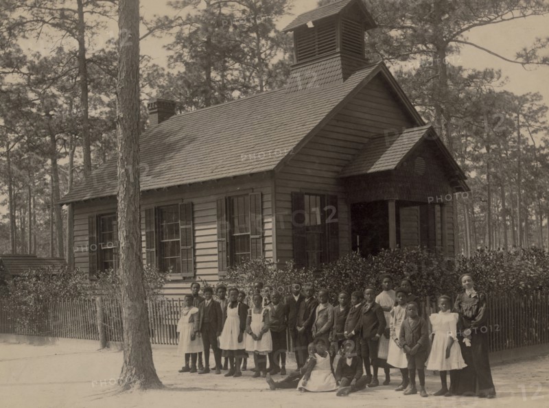 African American school children posed with their teacher outside a school, possibly in South Carolina