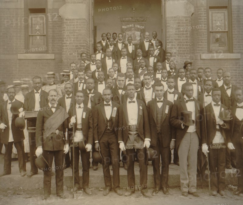 African American men posed at entrance to building, some with derbies and top hats, and banner labeled "Waiters Union" in Georgia
