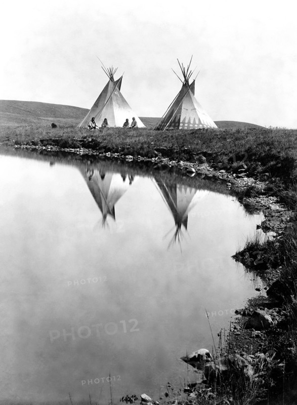 Edward S. Curits Native American Indians -  Two tepees reflected in water of pond, with four Piegan Indians seated circa 1910.