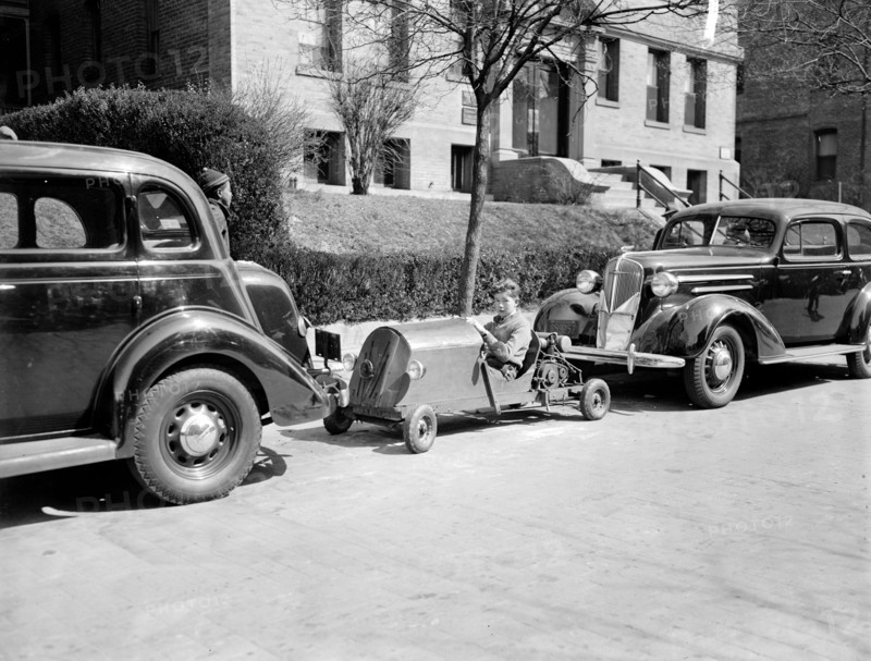 A boy occupies a parking spot in his home made go-kart circa 1937.