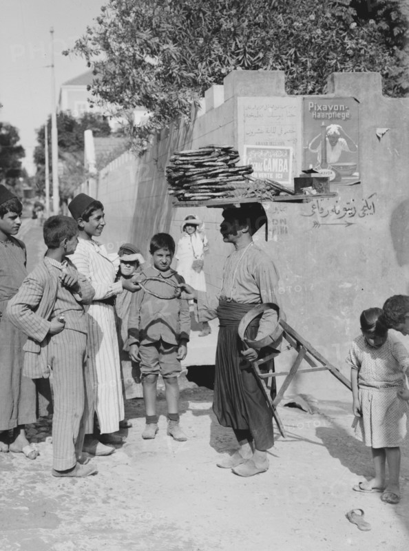 A man, street vendor, selling cakes in a Beirut street  to a crowd of young boys, man has cakes on top of his head ca. 1900