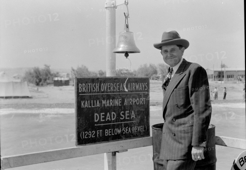 Man in a hat standing in front of a sign for the Dead Sea at Kallia Marine Airport  Aprril 5, 1946 ca.