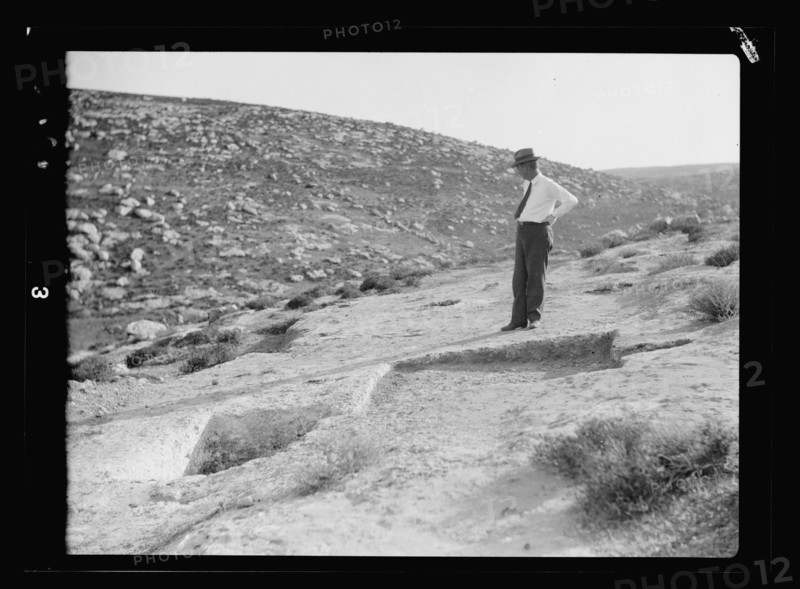 A man looks over an archelogical dig site and admires a primitive winepress showing treading-out vat ca. 1920