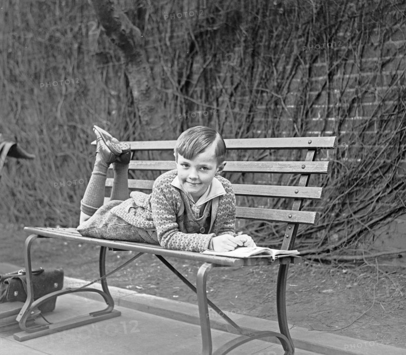 Child lying on outdoor bench with an open book ca. between 1909 and 1923