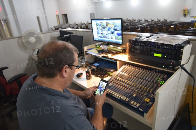 During the Covid-19 pandemic, some churches chose to record sermons to play on their websites and through streaming services to replace in person worship. Here, a deacon in the sound booth looks at his cell phone as a minister has his Sunday morning sermo