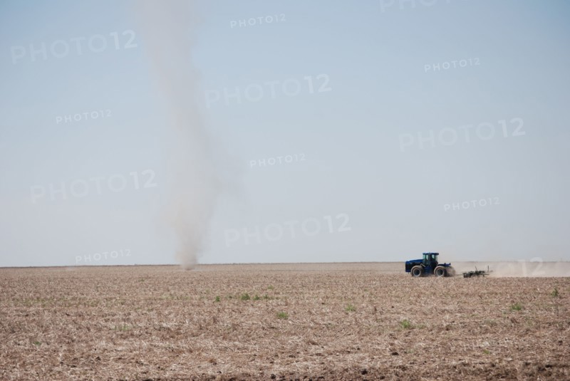 Farmer plowing a field on the Eastern Colorado plains, tractor facing left and driving toward a natural phenomena called a dust devil