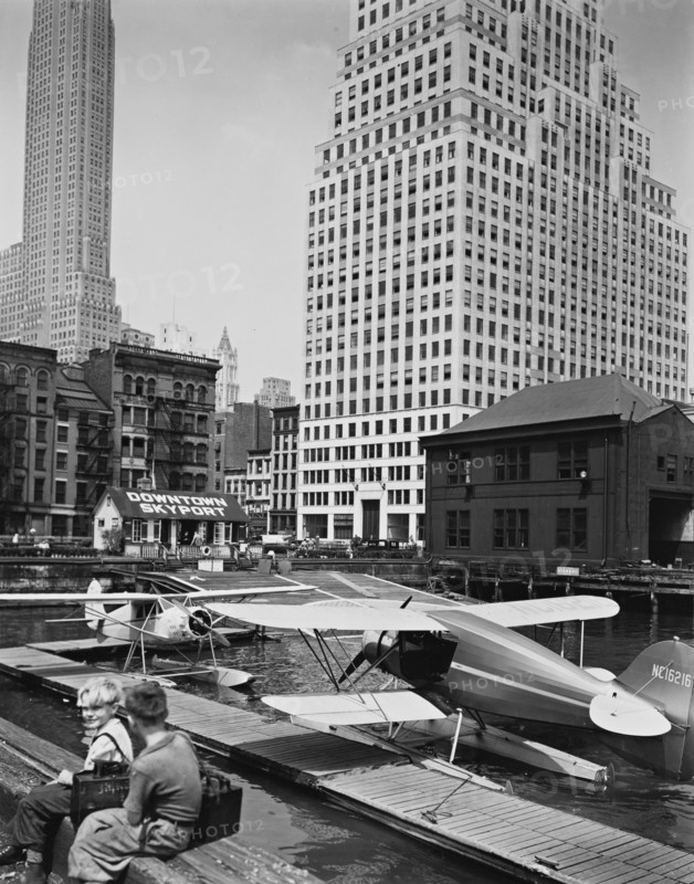 Two shoe-shine boys sit on edge of pier overlooking skyport, where seaplanes are moored; Manhattan office buildings beyond ca. 1936