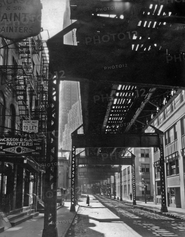 1930s New York City - Woman pauses below elevated train tracks, office buildings visible at left of tracks, painters shop in older building at left ca. 1936