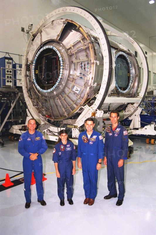 Members of the STS-88 crew pose with the Node 1 of the International Space Station in the high bay of the Space Station Processing Facility ca. 1997