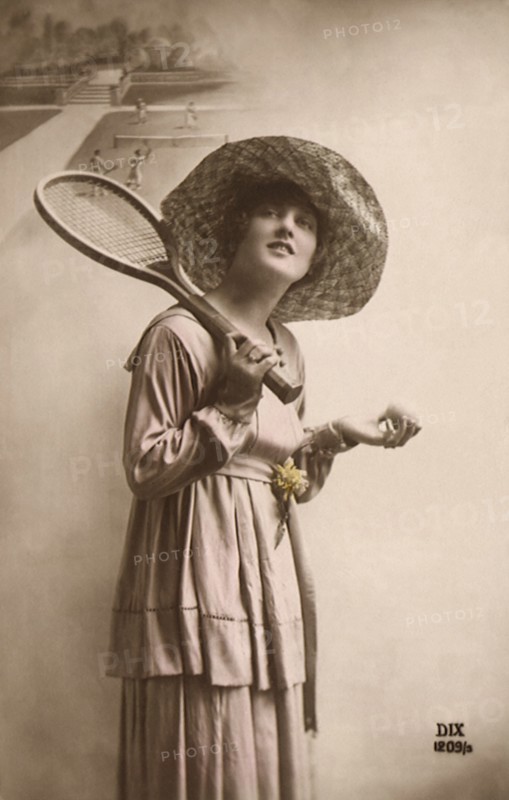 Woman in dress and hat with tennis racquet and ball.