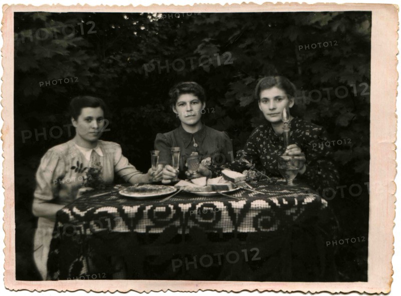 Three young women holding glasses of alcohol, sitting at a table covered with in garden.