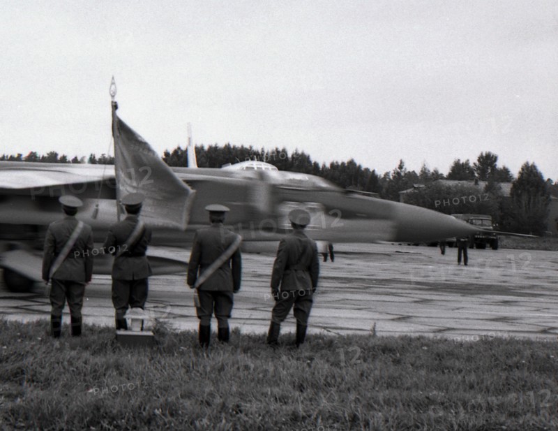 Four officers with regimental banner stand against the MiG-23 taxiing out for takeoff, air base Kubinka, USSR.