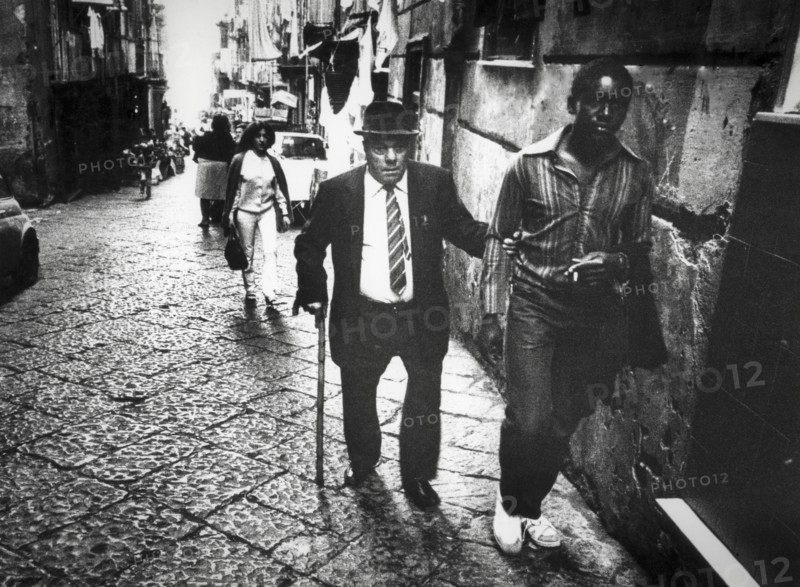 Elderly man and black man walking in a street of naples, 80's
