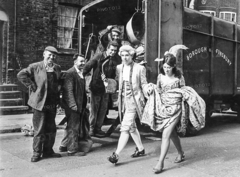 A couple of masked young people pass in front of a group of workers, london, england, uk. 1967