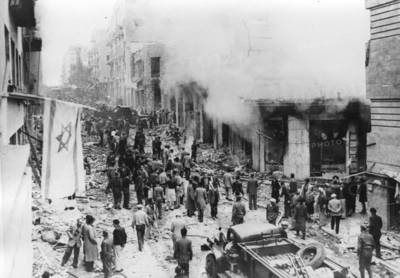 The Jewish Star of David flag hangs from its broken pole across from destroyed buildings and rubble.