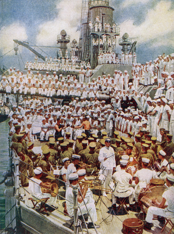 The caption reads: ""Pack up your troubles in your old sea bag. All work and no play was not permitted to make the American sailor a dull boy."" The image dates to 1922 and shows American military on a carrier at sea in World War I. In the foreground a band with a conductor. In the middle is a boxing matched watched by a number of sailors. The others are more sailors and crew watching the events.