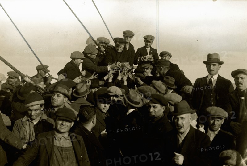 Group of emigrants around the chaplain on the foredeck of the Conte Verde