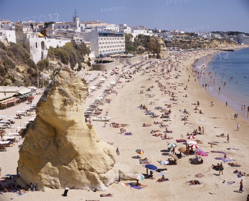PORTUGAL, Algarve, Albufeira, View along the beach from above with a large rock and sunbathers. Hotels and town beyond