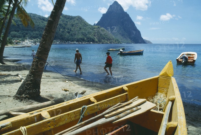WEST INDIES, St Lucia , Soufriere, People walking along beach with boats moored offshore and the Pitons beyond and prow of boat in imediate foreground.
