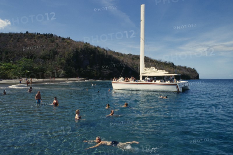 WEST INDIES, St Lucia, Transport, Catamaran tour.  Tourists swimming from moored boat with beach and tree covered headland behind.